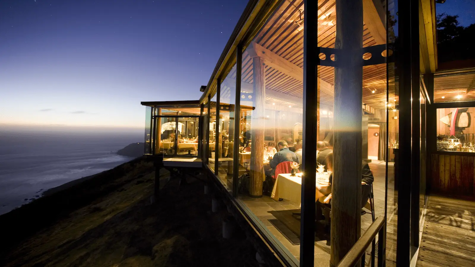 A glass-walled restaurant stands on the edge of a cliff at sunset, offering the best restaurant view on the Monterey Peninsula. The warm interior lights glow, creating a cozy ambiance. Diners inside enjoy their meals, while the expansive, darkening sky and sea create a serene backdrop.