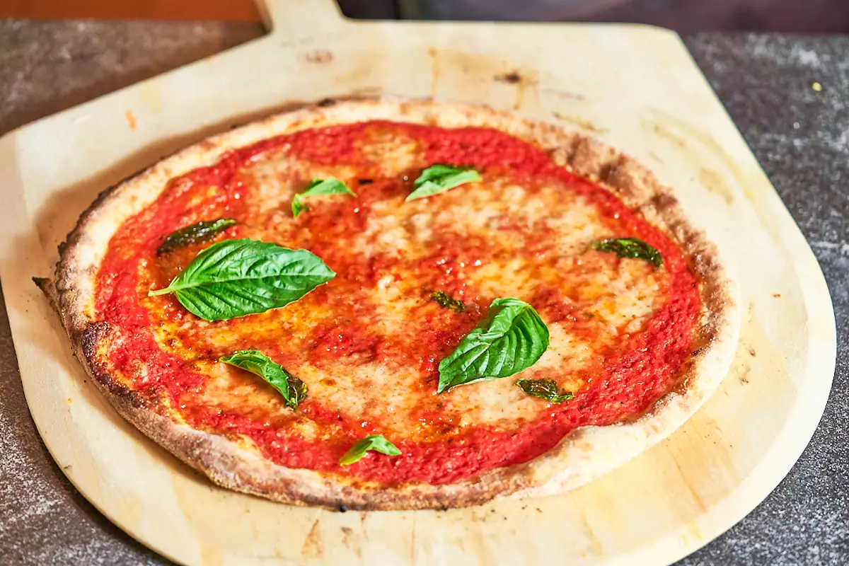 A freshly baked Margherita pizza displayed on a wooden pizza peel, perfect for New Year's Eve Celebrations 2023. The pizza has a golden brown crust, with a generous layer of tomato sauce, melted cheese, and scattered fresh basil leaves on top. The background shows a lightly floured surface.