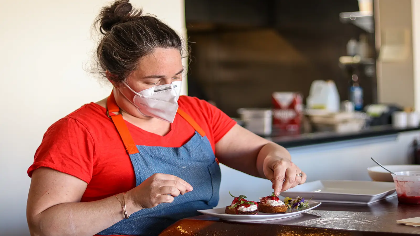 A person wearing a red shirt, blue apron, and face mask carefully places garnishes on plated dishes in a kitchen. The individual appears focused on the task, with various kitchen items visible in the background—a scene reminiscent of the best dinner spots in Sonoma County.