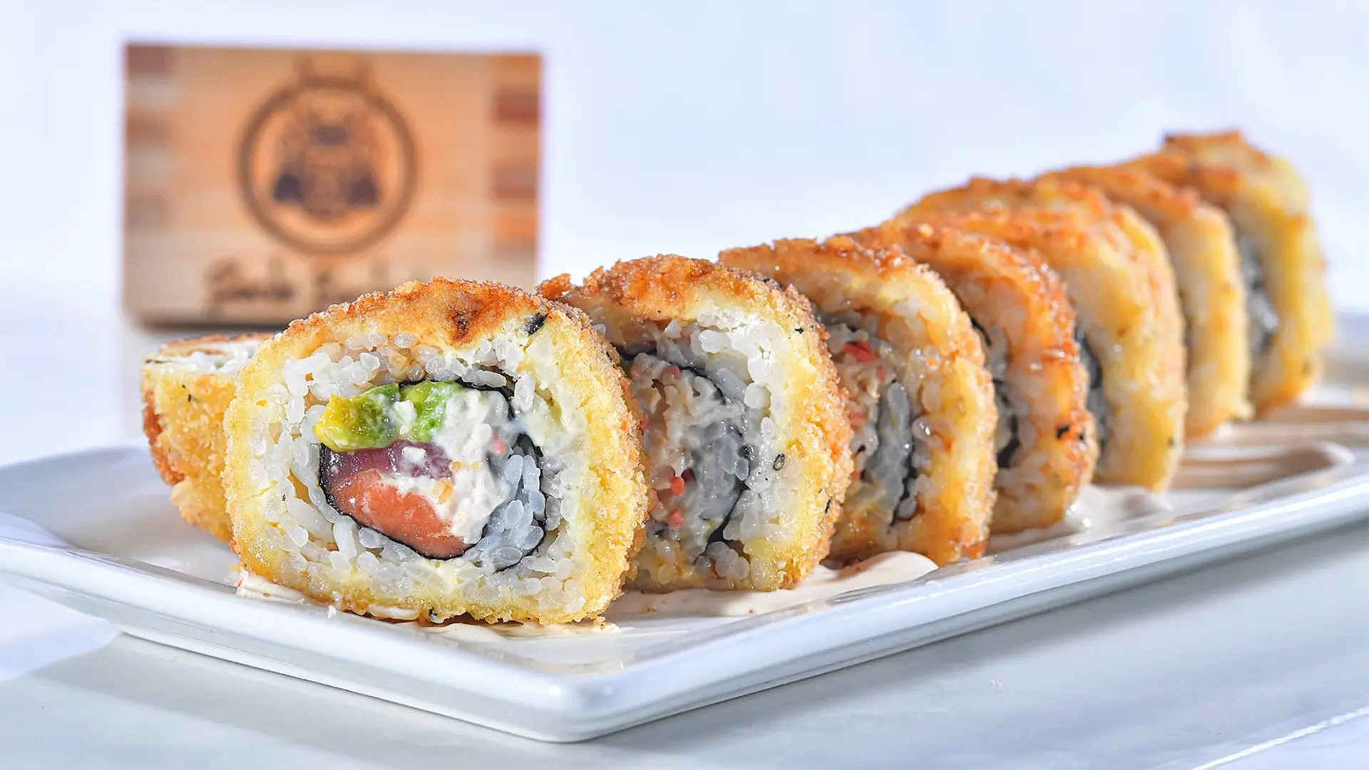 A close-up of a sushi plate featuring a roll of crispy, breaded sushi pieces lined up on a rectangular white plate. The sushi is filled with ingredients like avocado, cream cheese, and fish. In the background, partially visible but out of focus, is wooden wall decor reminiscent of the best sushi in Kauai.