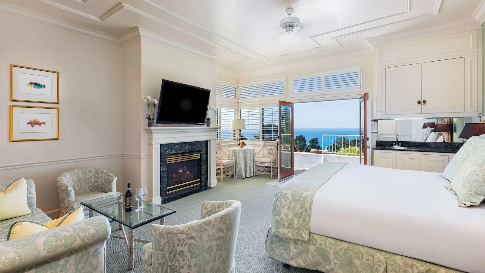 A spacious, elegant bedroom features a king-sized bed with light green and white bedding, armchairs, a fireplace with a TV above it, and large windows with a patio door opening to an ocean view. The walls are adorned with framed artwork and cream-colored paneling—truly an ideal place when considering where to stay on the Monterey Peninsula.