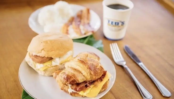 A breakfast meal is served on a wooden table. It includes a croissant sandwich with bacon, egg, and cheese, a biscuit sandwich, a plate with rice and bacon, and a cup of coffee with "TED'S" printed on it. A fork and knife are placed next to the plate—truly the best breakfast North Shore has to offer.