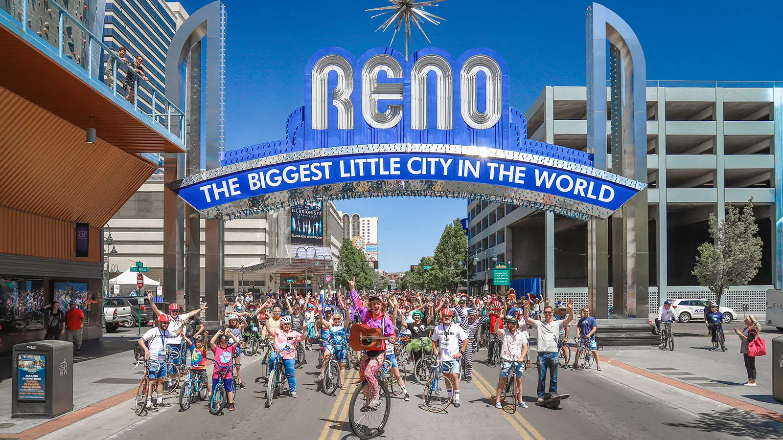 A large group of cyclists participate in a colorful bike parade under the iconic Reno arch that reads "Reno: The Biggest Little City in the World." The sky is clear, buildings line both sides of the street, and spectators watch. It's one of the best things to do in Tahoe June events.