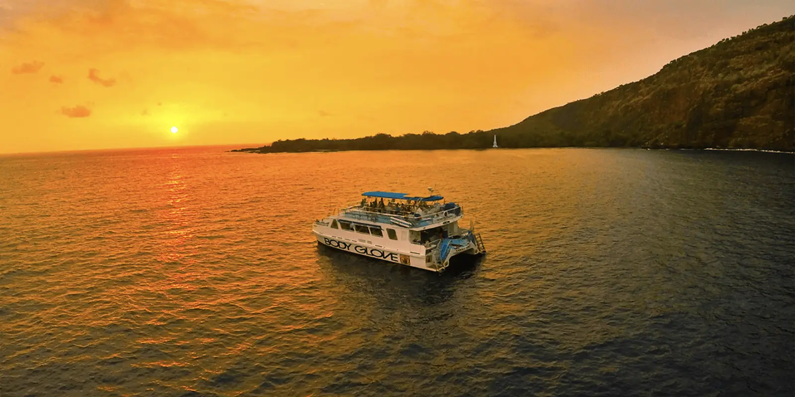 A boat with "Body Glove" written on its side floats on a calm ocean at sunset, captured perfectly by one of the Top Big Island Sunset Tours. The sky is ablaze with vibrant orange hues, and the sun is near the horizon. A mountainous coastline is visible in the background, casting a serene reflection on the water.