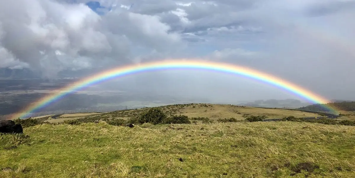A vibrant rainbow arches across a mostly cloudy sky over a green hilly landscape, reminiscent of the vista you'll encounter on the best island tour Maui offers. The terrain, adorned with sparse vegetation and a winding road, seamlessly blends into the natural scenery.