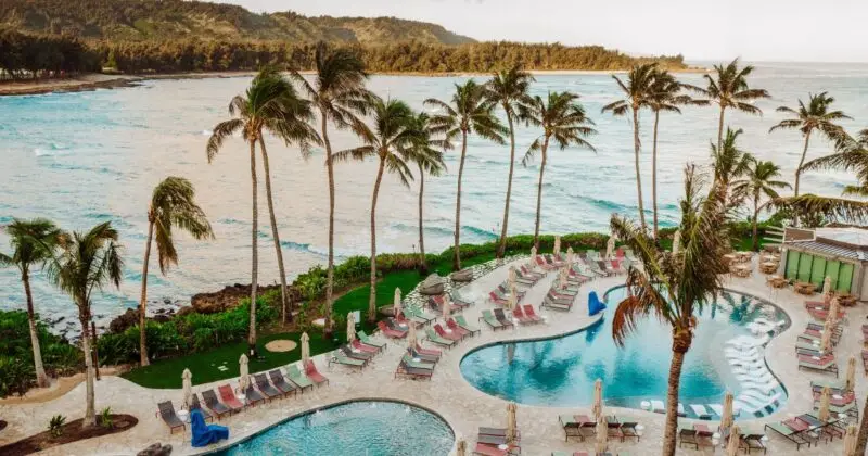 A scenic view of a tropical resort featuring two curvy hotel pools surrounded by several lounge chairs, umbrellas, and palm trees. The resort overlooks a beautiful coastline with gentle waves and a backdrop of lush green hills. This idyllic setting perfectly captures the essence of paradise.