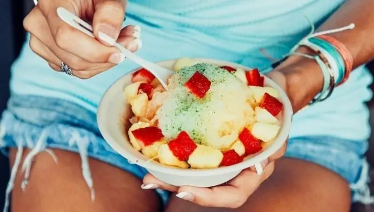A person holding a bowl of the best shave ice in Waikiki, topped with green syrup and surrounded by chunks of red and yellow fruit. The person is dressed in a light blue top and denim shorts, with colorful bracelets on their wrist, using a white spoon to scoop the delightful dessert.
