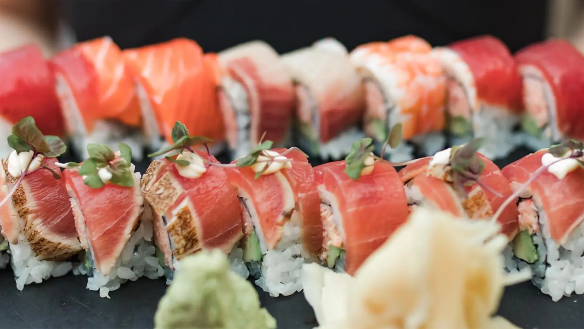 A close-up of a beautifully arranged sushi platter, perfect for dinner in Paia and Hana Maui, featuring various types of sushi rolls, including slices of tuna, salmon, and other fish topped with decorative microgreens. Wasabi and pickled ginger are placed in the foreground.