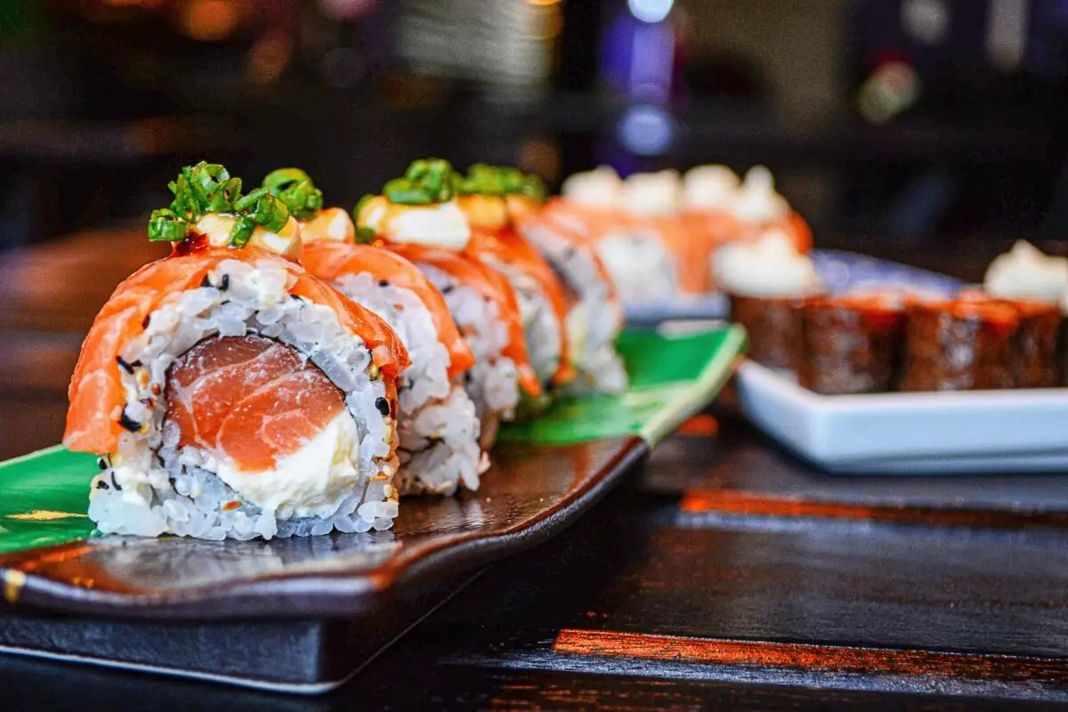 A close-up of a sushi roll topped with diced green onions, featuring slices of salmon, rice, and cream cheese on a dark rectangular platter. In the background, another dish of sushi rolls from the best sushi spots in Princeville is slightly out of focus.