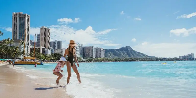 A woman and a child walk along a sandy beach with their feet in the water. The woman, wearing a hat and swimsuit, strolls alongside the child in a floral outfit and hat. Tall buildings and a mountain are visible under Waikiki's clear blue sky, with an outrigger canoe in the distance.