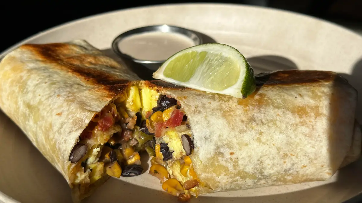 A close-up of one of the best breakfast burritos on the Kohala Coast, cut in half on a white plate. The burrito contains beans, rice, corn, tomatoes, and scrambled eggs. Topped with a lime wedge, it is served with a small container of light-colored dipping sauce. The dish is well-lit with natural light.
