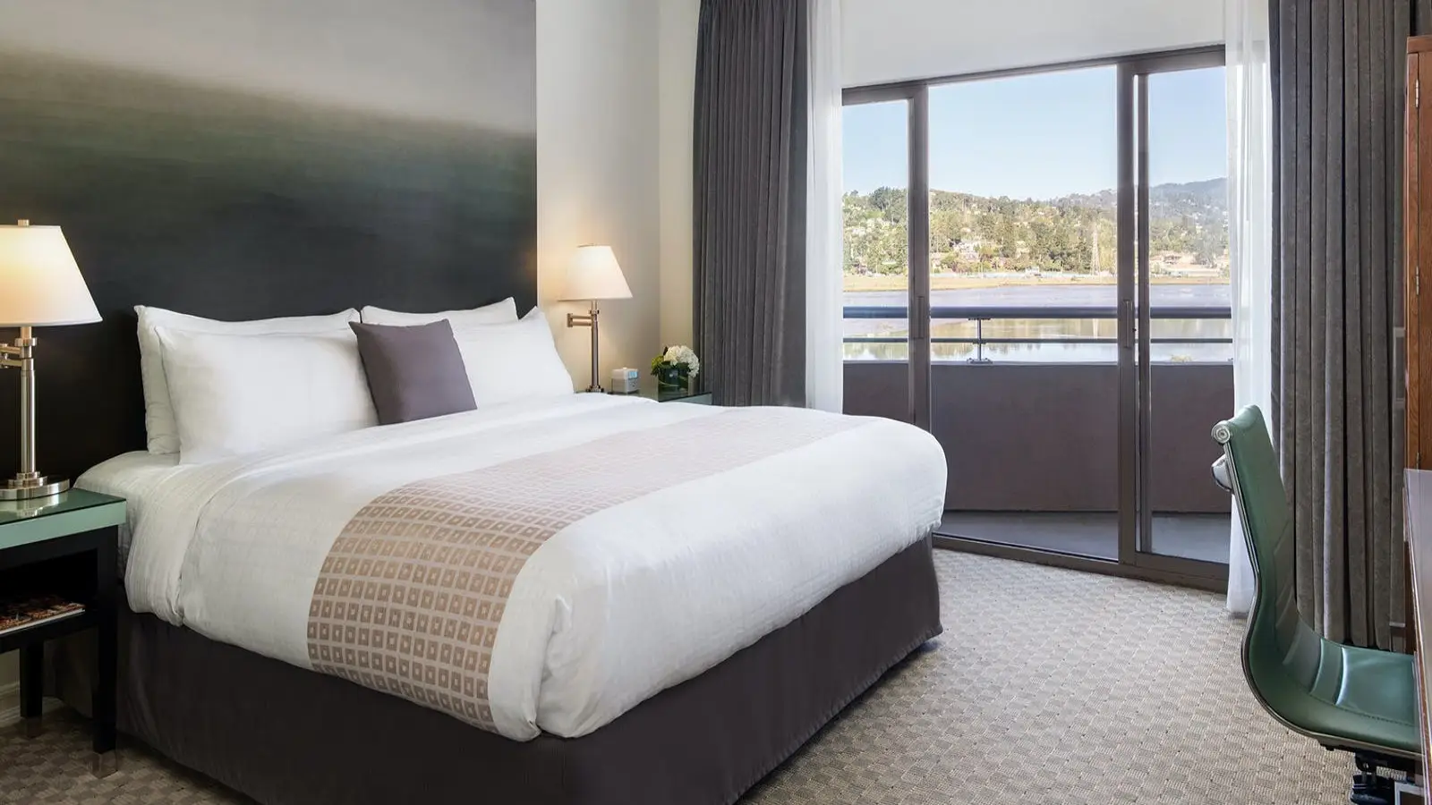 A modern hotel room in one of the best group hotels North Bay has to offer, featuring a neatly made king-size bed with white linens and a decorative throw. The room includes nightstands with lamps, a green desk with chair, and a large sliding glass door opening to a balcony with a scenic view.