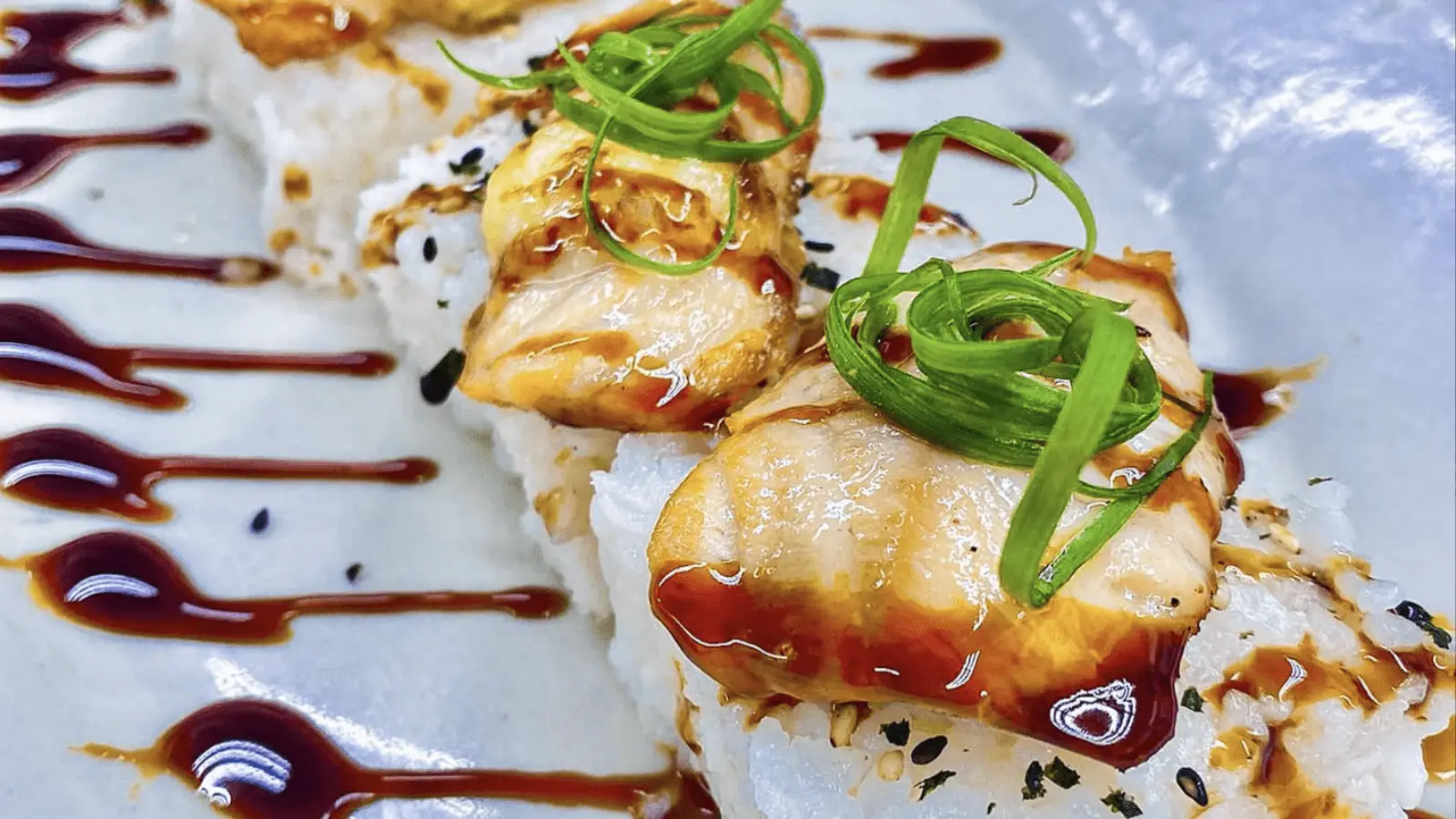 Close-up of sushi rolls topped with seared fish slices, drizzled with a dark sauce, and garnished with thin green onion strips. The sushi is plated on a white dish with additional sauce artistically streaked across the plate, making it one of the best dinner options in Ko Olina.
