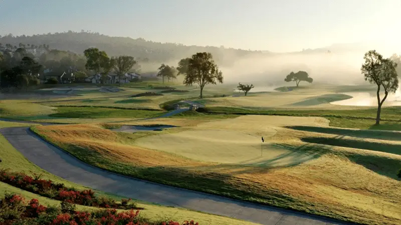 A picturesque golf course at sunrise, with lush green fairways perfect for golfing, a winding path, and a flagstick marking a hole. Trees and shrubs are scattered throughout the landscape, with mist hanging in the air and distant hills partly obscured by fog.