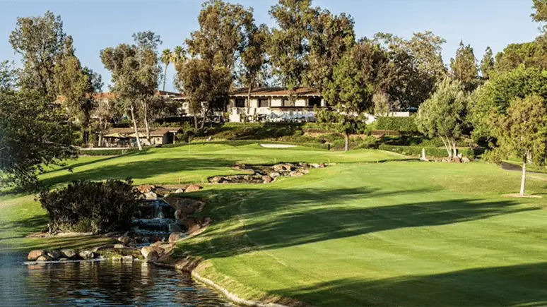 A scenic view of one of the best golf courses in the Bay Area, featuring lush green fairways, a water hazard with a small waterfall, and surrounding tall trees. In the background, there's a clubhouse with outdoor seating. The sky is clear and blue. Perfect for a day of golfing or exploring things to do in San Diego.
