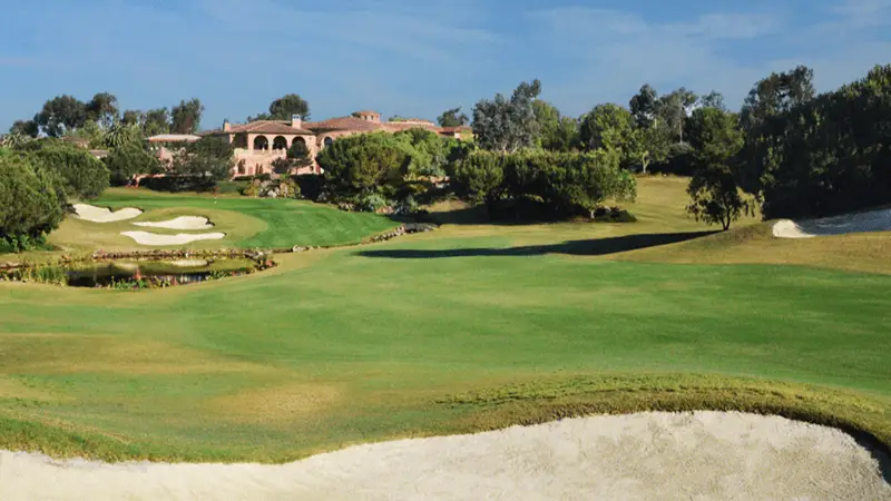 A scenic golf course in San Diego with a sprawling green fairway, sand bunkers, and a pond. A large, elegant house with arched windows and a terracotta roof is visible in the background, surrounded by trees and landscaped greenery under a clear blue sky—perfect for golfing enthusiasts.
