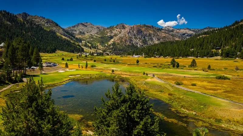 A scenic landscape featuring a lush green golf course, perfect for golfing, surrounded by tall trees and a small pond in the foreground. The backdrop includes rugged mountains under a clear blue sky, with scattered clouds enhancing the natural beauty of one of the best golf courses Tahoe has to offer.