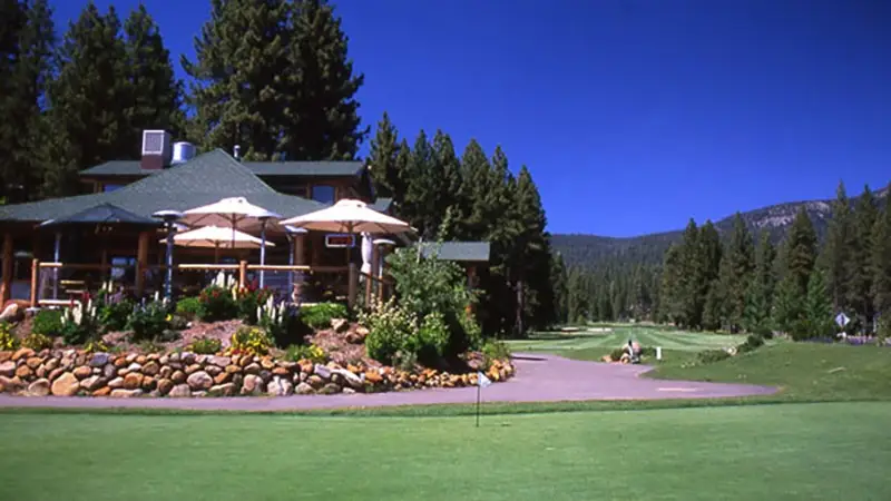 A golf course is pictured with a clubhouse on the left featuring a patio shaded by large umbrellas. The building is surrounded by lush green trees and landscaping. The clear blue sky and mountainous backdrop enhance the serene outdoor setting, making it one of the best golf courses Tahoe has to offer.