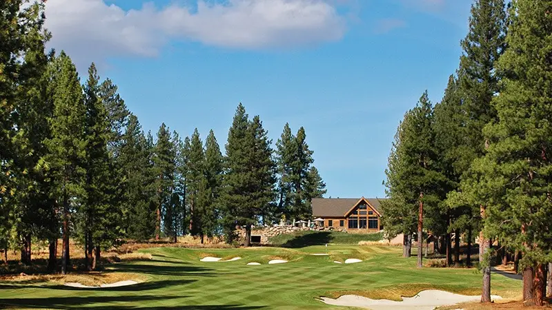 A scenic golf course featuring a well-maintained green with several sand bunkers. Tall pine trees border both sides, and a rustic, wooden clubhouse sits in the background under a clear blue sky with a few clouds, making it one of the best golf courses Tahoe has to offer.