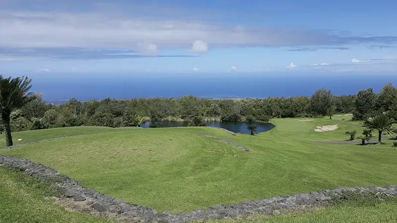 A scenic view of one of the best golf courses on the Big Island features a lush green expanse with a small pond. In the background, trees and a clear blue sky with a few clouds extend to the horizon where the sky meets the blue ocean. A stone wall borders a section of this pristine course.