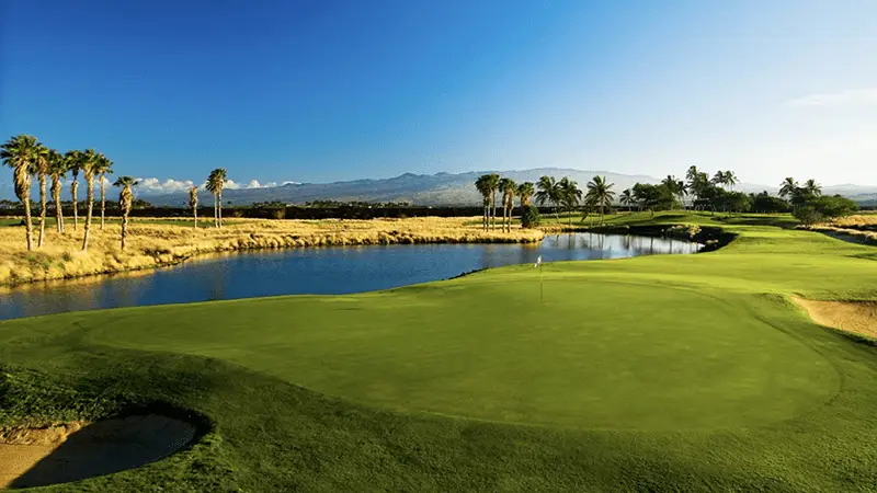 A scenic golf course on the Big Island features a lush, green fairway with a flagstick planted in the hole. A clear blue pond runs alongside, bordered by palm trees and sand bunkers. The sky is clear with a few scattered clouds, and distant mountains are visible—one of the best golf courses you'll find.