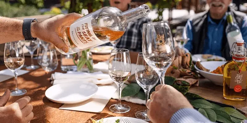 A hand pours rosé wine from a bottle labeled "Finn's Bay Sand Beach" into a wine glass at an outdoor dining table, perfect for local getaways. The table is set with plates of food, wine glasses, and a few bottles, with people seated around it enjoying the meal.