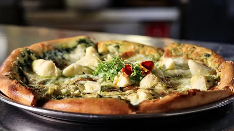 A delicious pizza topped with artichoke hearts, greens, and other ingredients sits on a silver tray. The crust is golden-brown and the toppings look fresh and flavorful. Enjoy the best lunch on the Kohala Coast as you savor every bite in a kitchen setting slightly out of focus.