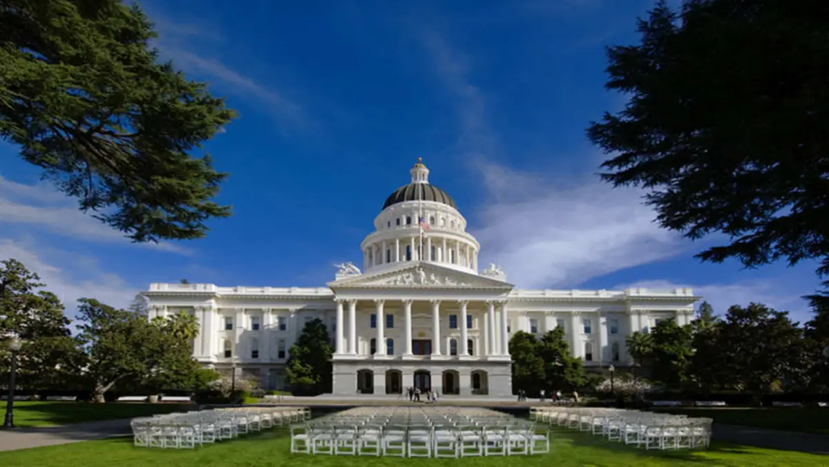 The image shows a large, white, neoclassical building with a prominent dome, columns, and steps, likely one of the best places to visit in Sacramento. In front are rows of white chairs arranged on a lawn, surrounded by trees, under a clear blue sky.