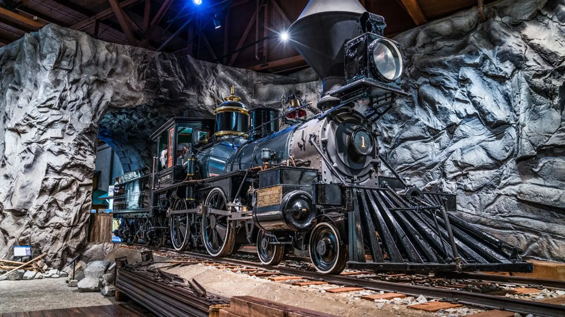 A vintage steam locomotive is displayed inside an exhibit that simulates a rocky tunnel. The train, black with intricate brass fittings and prominent front lights, stands on track rails amidst wooden beams and blue accent lighting overhead. It's one of the best places to visit while exploring Los Angeles.