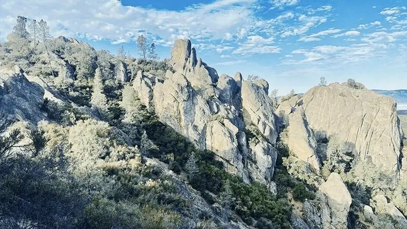 A rugged mountain landscape featuring dramatic rock formations under a bright blue sky with scattered white clouds, reminiscent of the terrain surrounding Central California missions. Sparse vegetation, including trees and shrubs, grows among the rocky terrain, showcasing a combination of cliffs, peaks, and valleys.