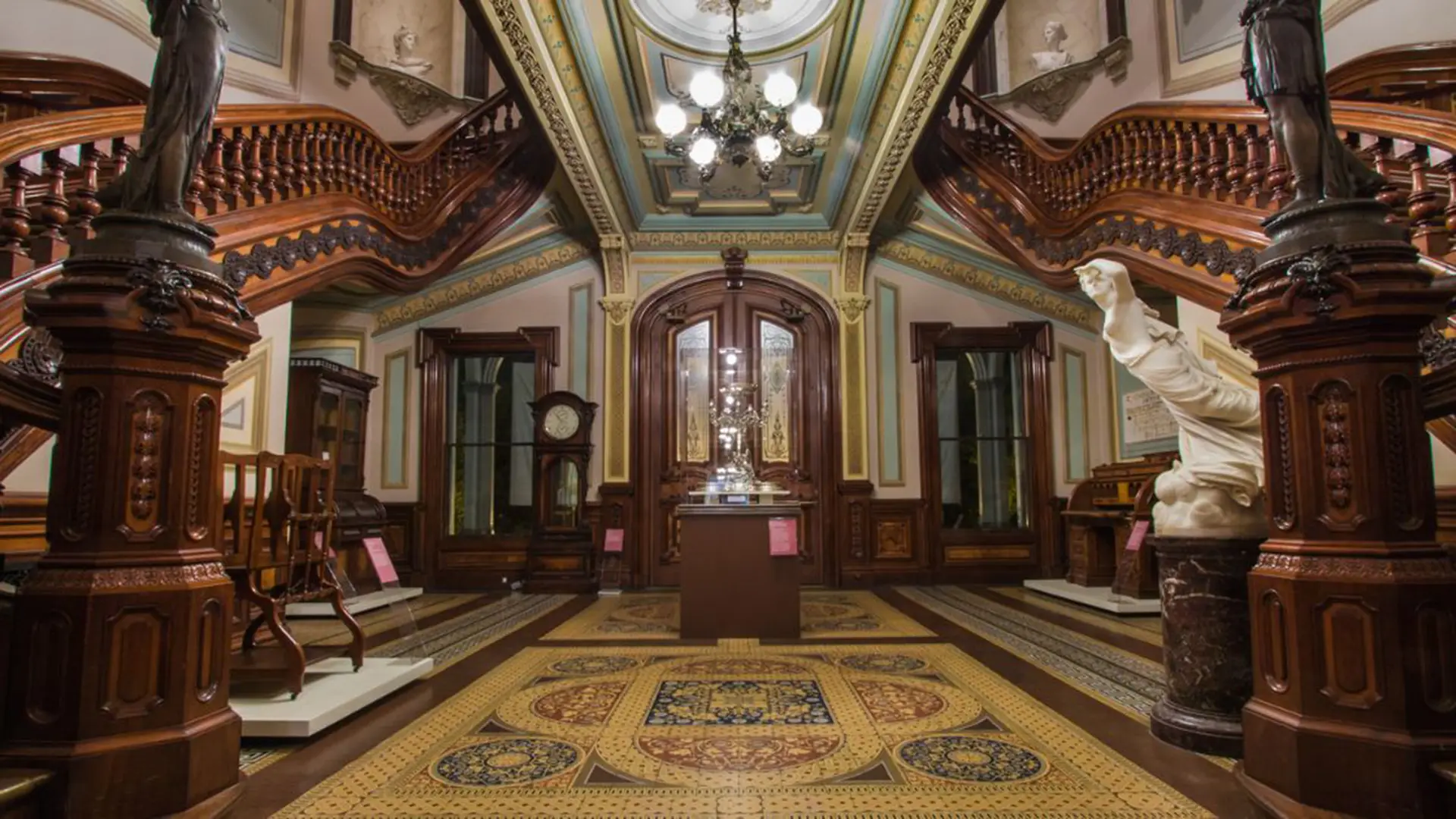 A richly decorated room features ornate wooden furniture, a central glass display case, grand chandeliers, and intricate ceiling designs. The room has detailed wood paneling, vintage carpeting, and several statues and artifacts on display—one of the best places to visit in Sacramento for history buffs.