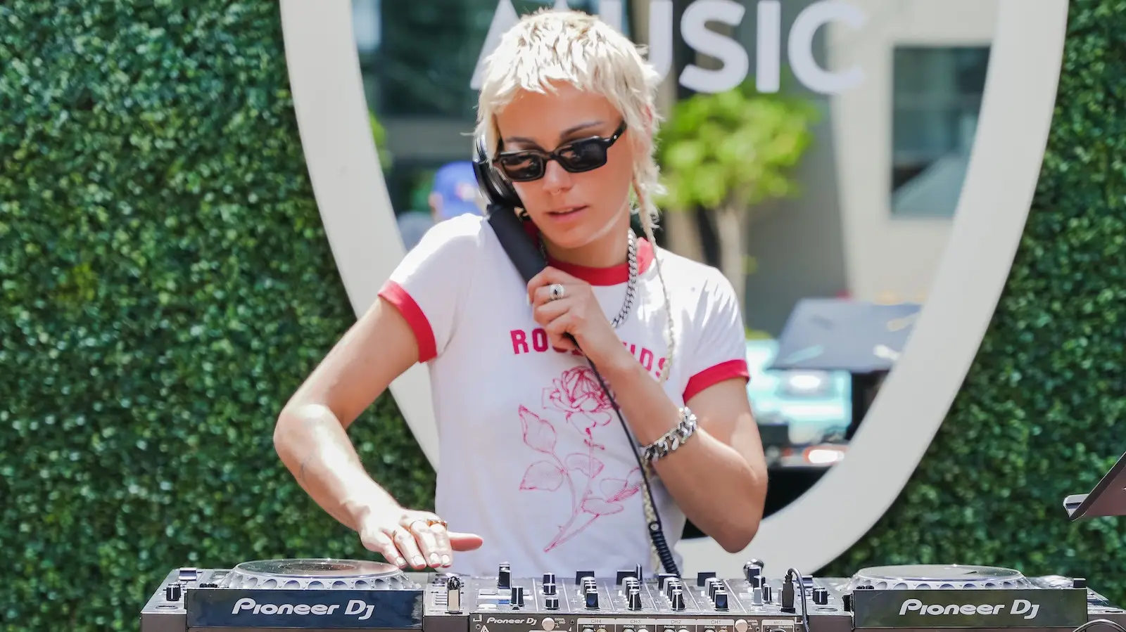 A person with short blonde hair wearing sunglasses, a white t-shirt with red trim, and casual accessories is DJing on Pioneer DJ equipment outdoors at Hotel Ziggy. They are holding a headphone to their ear with one hand and adjusting the mixer with the other.