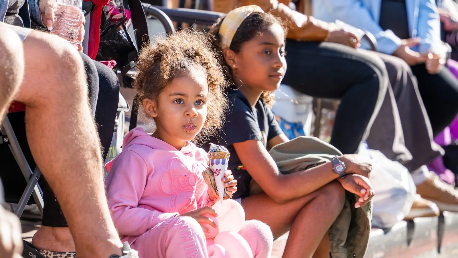 Two young children are sitting outdoors in the coastal town of Tiburon, watching something off-camera. The younger child, wearing a pink tracksuit, holds a snack, while the older child, wearing a black shirt and a headband, is seated next to her. Other people are around them, partially visible.