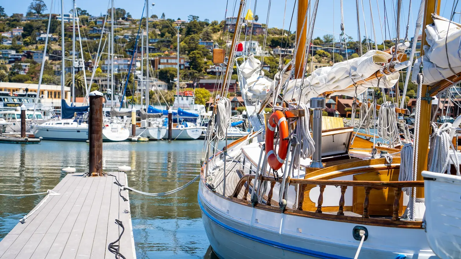 A sunny marina scene in Tiburon features a wooden sailboat moored to a dock. Behind the sailboat, more boats are docked in the bustling harbor. The hillside in the background is dotted with houses and lush greenery, while reflections of the boats shimmer on the calm water.