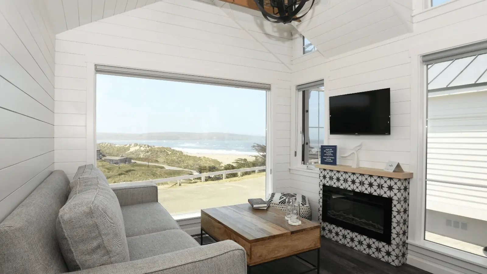 A cozy, modern living room in one of the best group hotels North Bay has to offer, with a large window offering a stunning view of the beach and ocean. The room features a gray sofa, wooden coffee table, TV mounted above a patterned fireplace, and white shiplap walls. Decor is minimal and contemporary.