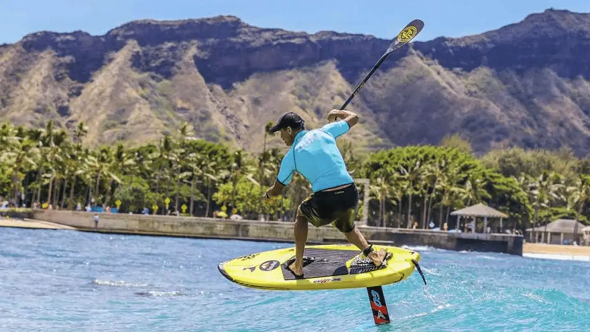 A person is riding a hydrofoil board over blue ocean water near a tropical shoreline with palm trees and a mountainous backdrop under a clear blue sky. Embracing the spirit of Hawaiian holidays, the rider is wearing a hat, a blue shirt, and black shorts, and holding a paddle while balancing on the board.