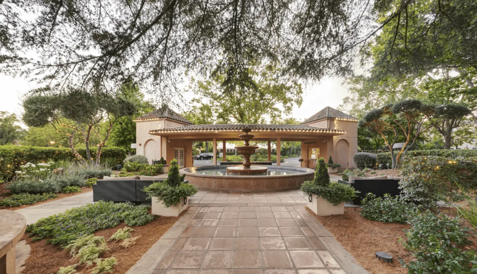 A paved walkway leads to a large, ornate fountain surrounded by well-manicured greenery. The fountain is situated in front of a stucco building with a tiled roof, characteristic of a wine country getaway. The area is adorned with small lights and shaded by overhanging tree branches.