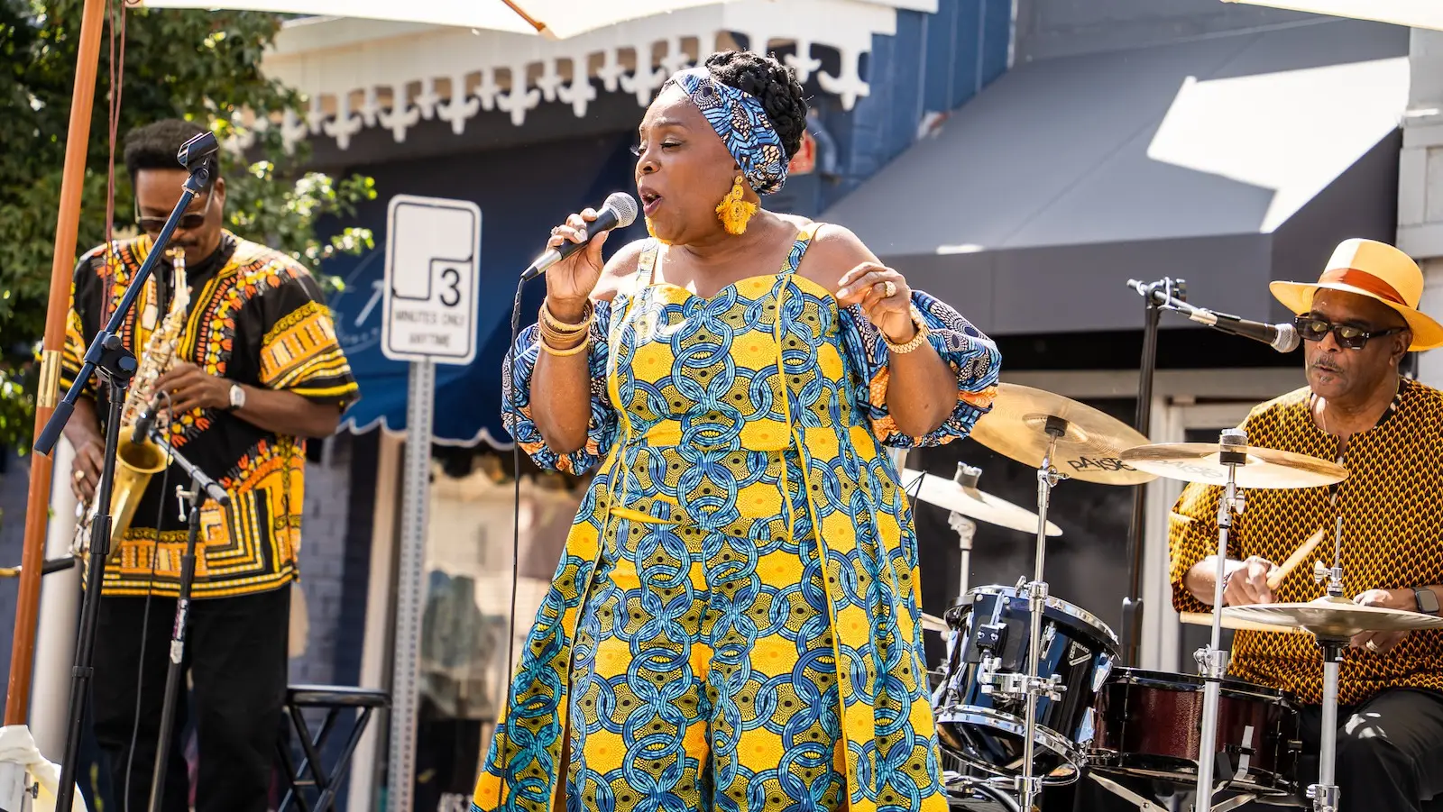 A woman wearing a vibrant blue and yellow patterned dress and headwrap sings into a microphone at an outdoor performance. A man on her left plays a trumpet, while a drummer with sunglasses and a yellow hat is on her right. They are under large umbrellas, reminiscent of the lively street scenes in Tiburon.