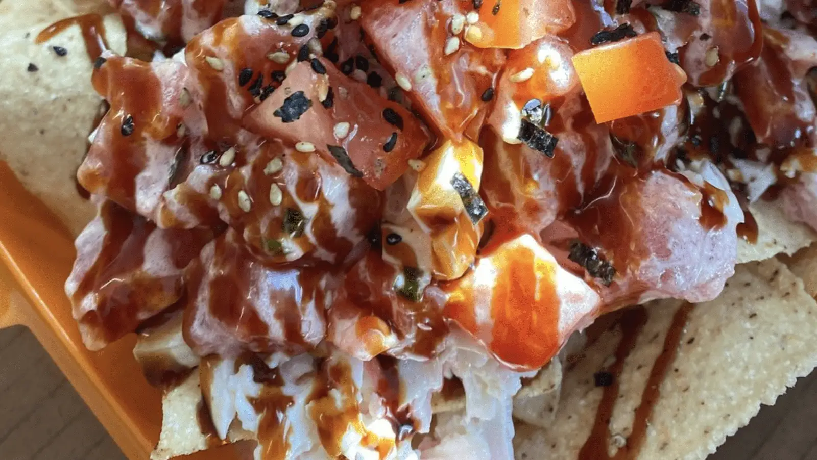 A close-up of a dish featuring the best poke on the Big Island, showcasing diced raw fish garnished with diced tomatoes, a drizzle of dark sauce, and a sprinkle of sesame seeds and seaweed flakes, all served on a bed of tortilla chips.