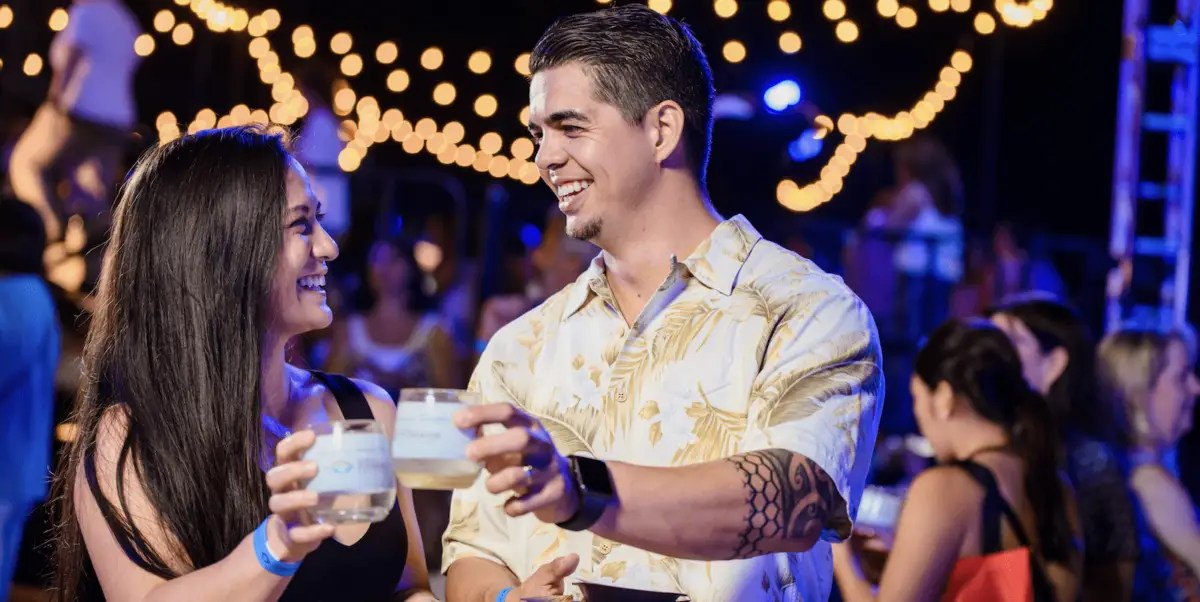 Two people are smiling and clinking glasses at a lively outdoor event. They are surrounded by a crowd, with string lights creating a festive atmosphere in the background. The person on the right, who has a tattoo on their forearm, is wearing a patterned shirt typical of Annual Events Waikiki.