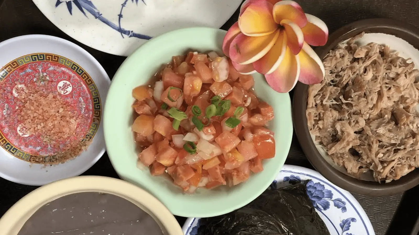 A selection of traditional dishes defining Hawaiian culture is displayed, including a bowl of diced tomatoes garnished with chopped green onions, a plate of shredded pork, a bowl of pink salt, a dish with leafy greens, and a bowl of smooth, purple dessert adorned with decorative flowers.