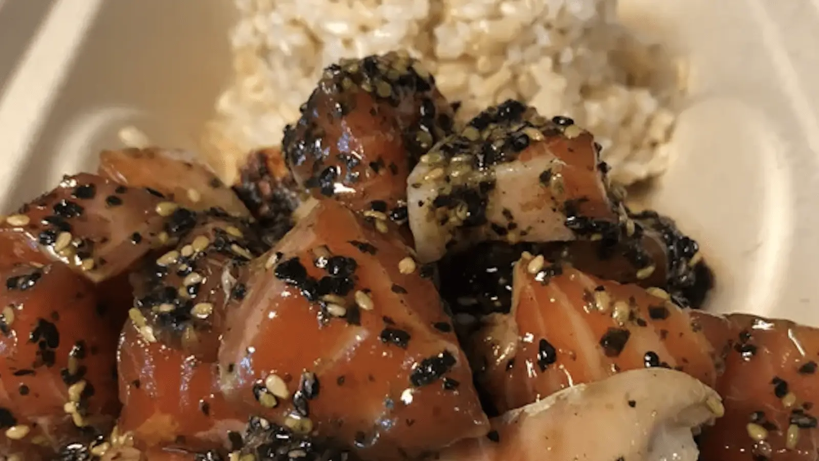 A meal featuring cubed raw fish seasoned with sesame seeds and herbs, reminiscent of the best poke on the Big Island, served with a side of brown rice. The dish is neatly presented in a takeout container.