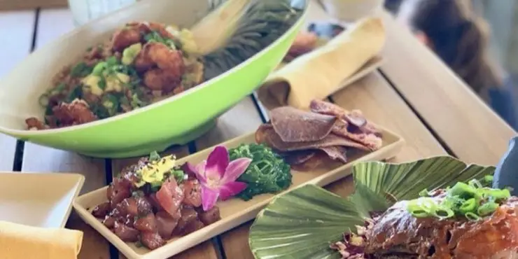 A table with plates of colorful food, including a bowl of fried chicken garnished with green onions, a rectangular plate with the best poke on the Big Island featuring diced raw fish, seaweed salad, taro chips, and an edible flower, and a banana leaf-wrapped dish set atop a wooden surface.