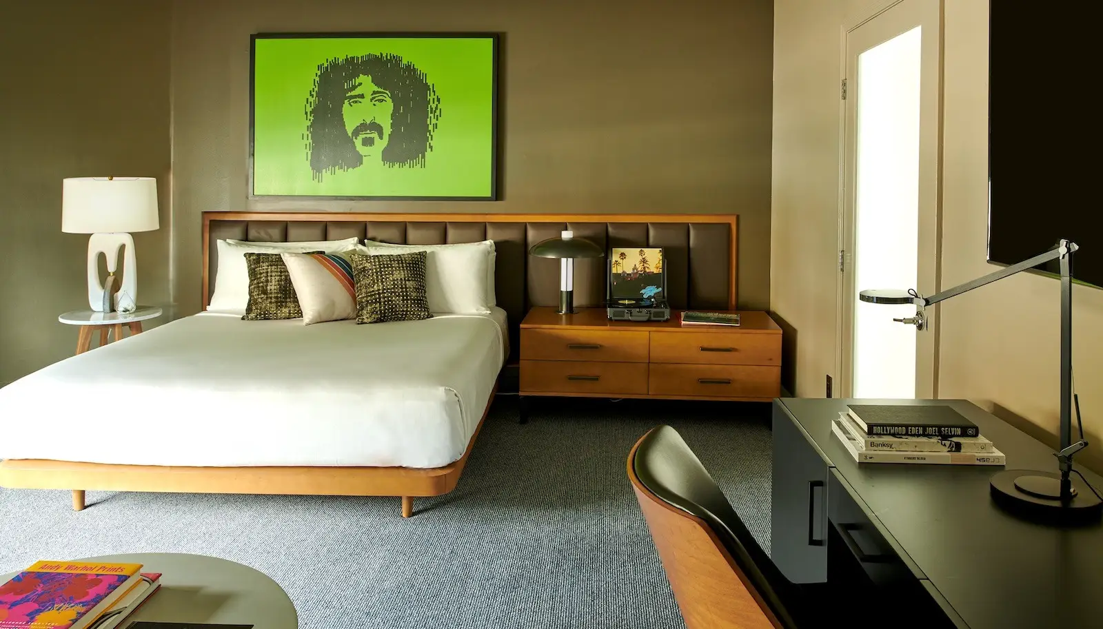 A modern bedroom at Hotel Ziggy features a large bed with multiple pillows, a green artwork above the headboard, a side table with a lamp and book, and a wooden desk with a chair and sleek desk lamp. The room has neutral tones with green accents and abundant natural light.