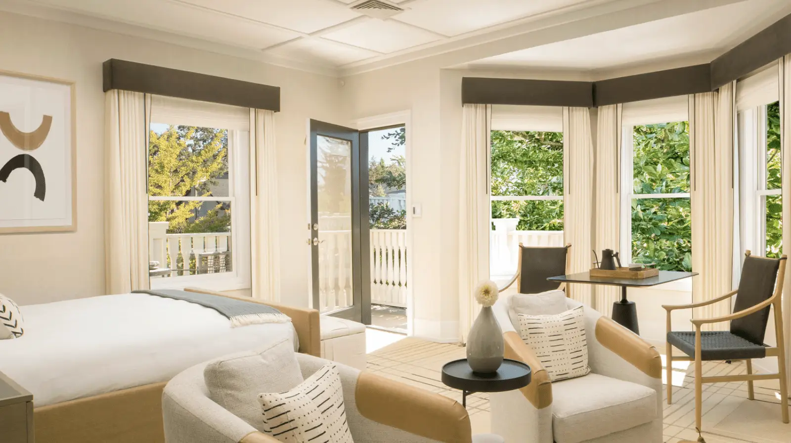 Bright, modern Sonoma bedroom with light walls and large windows. The room features a bed, two chairs with side tables, a small round table with two chairs, and a balcony with white railings. Greenery is visible outside the windows and the balcony door.