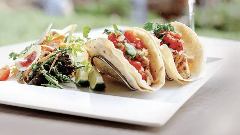 A close-up of a white plate featuring three tacos filled with vegetables and garnished with cilantro. On the left side is a small salad with mixed greens, tomatoes, and thinly sliced radishes. Lime wedges are placed behind the tacos—truly the best dinner Ko Olina has to offer.