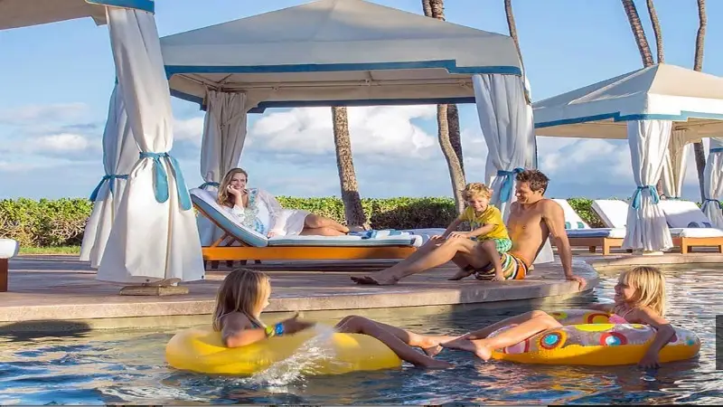 A family enjoys a sunny day by the pool. A woman lounges in a poolside cabana while a man sits on the edge of the pool, engaging with two children floating in inflatable pool toys. Palm trees and more cabanas are visible in the background, creating an idyllic scene for local getaways.