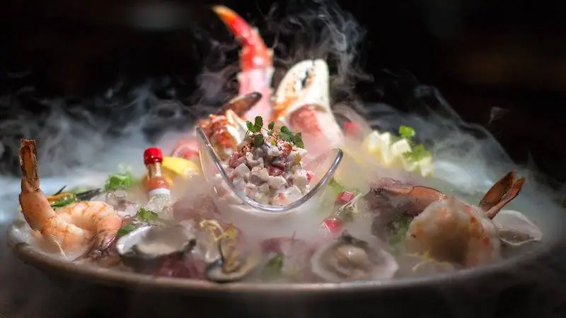 A luxurious seafood platter, perfect for the best dinner in Ko Olina, features an assortment of fresh shrimp, crab legs, oysters, and clams beautifully presented on a bed of ice with a fog effect. A glass bowl of ceviche garnished with herbs is prominently placed in the center.