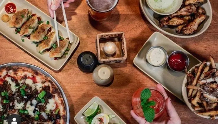 A wooden table is filled with various dishes, including a pizza, dumplings, chicken wings, fries, and a cocktail garnished with mint. A hand uses chopsticks to pick up a dumpling while another holds a red cocktail. There are sauces and a cup of beer nearby—truly the best dinner in Ko Olina.