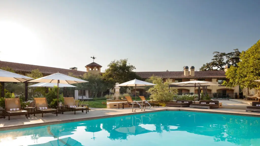 A luxurious outdoor pool area at a resort, perfect for local getaways, features several lounge chairs under umbrellas arranged around a pristine pool. The backdrop includes lush greenery and a large building with a red-tiled roof. The sky is clear, indicating a sunny day.